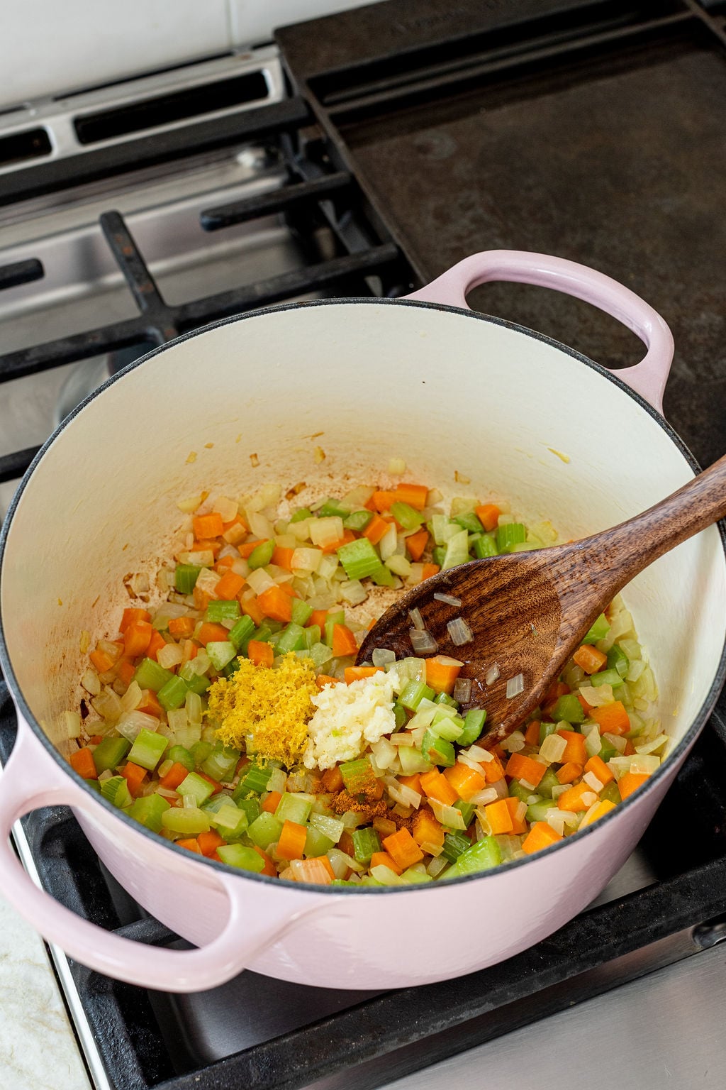 Diced carrots, celery, and onions are being saut&eacute;ed in a pink Dutch oven, with minced garlic and lemon zest added, stirred by a wooden spoon on a stovetop.