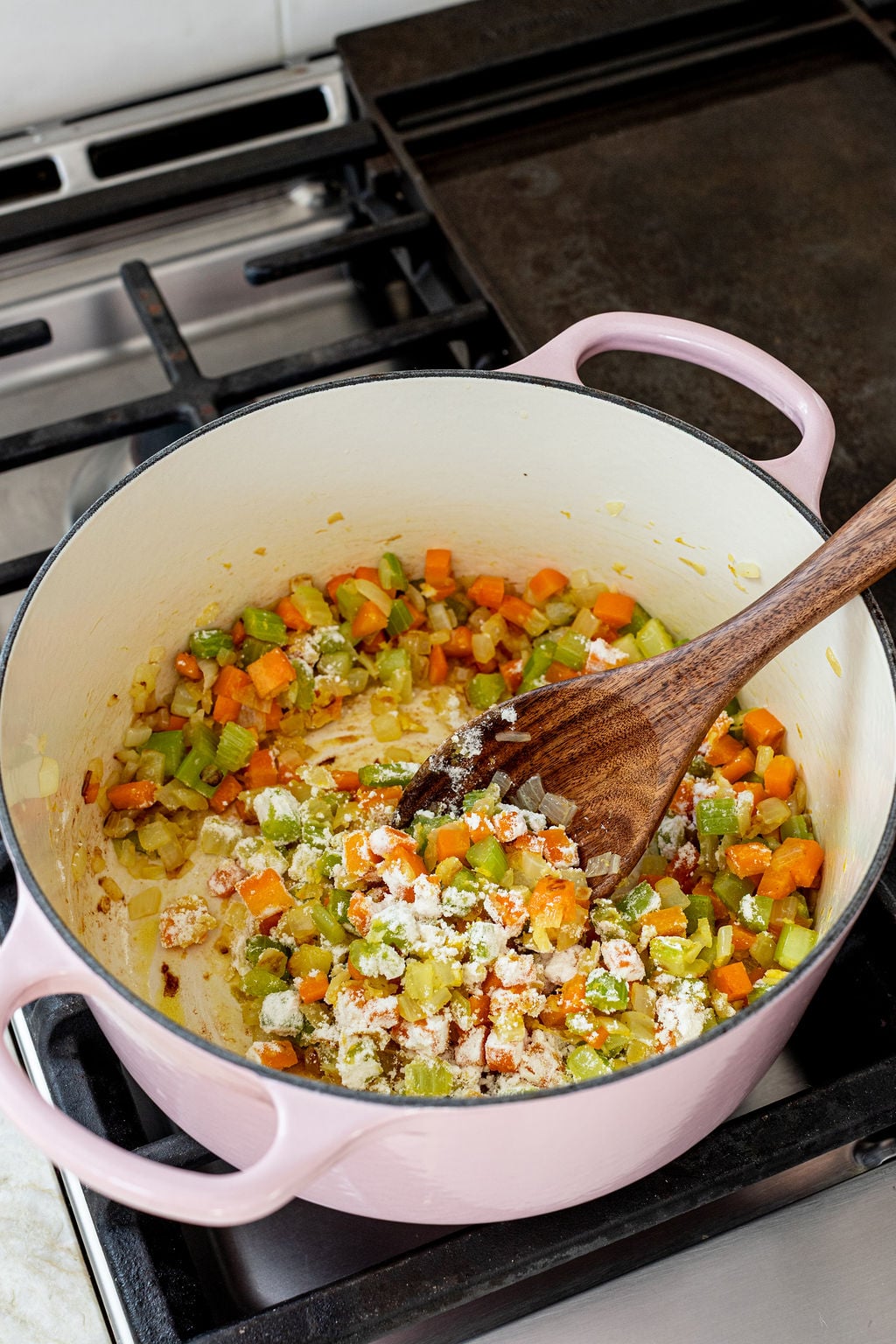 A pink Dutch oven on a stovetop contains chopped carrots, celery, onions, and flour being stirred with a wooden spoon.
