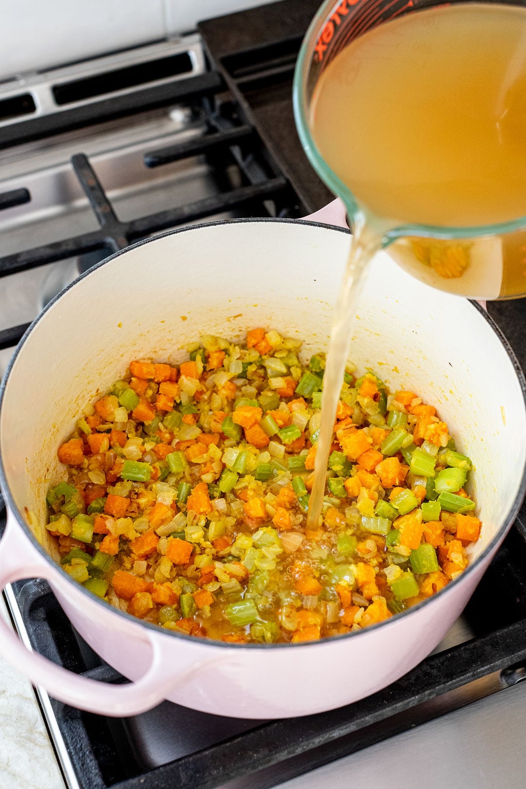 Chopped carrots, celery, and onions are saut&eacute;ing in a pink pot on the stove as vegetable broth is poured in from a measuring cup.