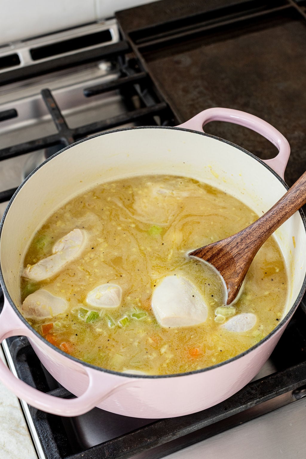 A pink Dutch oven on a stove contains soup with several pieces of chicken, vegetables, and broth; a wooden spoon rests inside the pot.