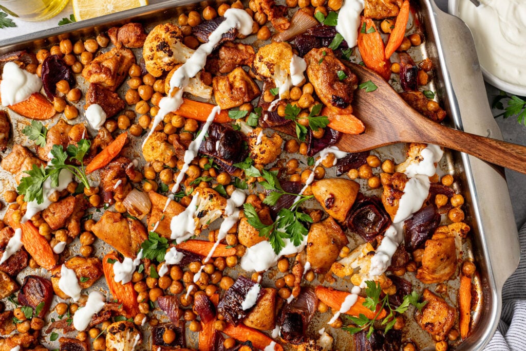 A sheet pan filled with roasted vegetables and chicken thighs drizzled with creamy yogurt sauce.