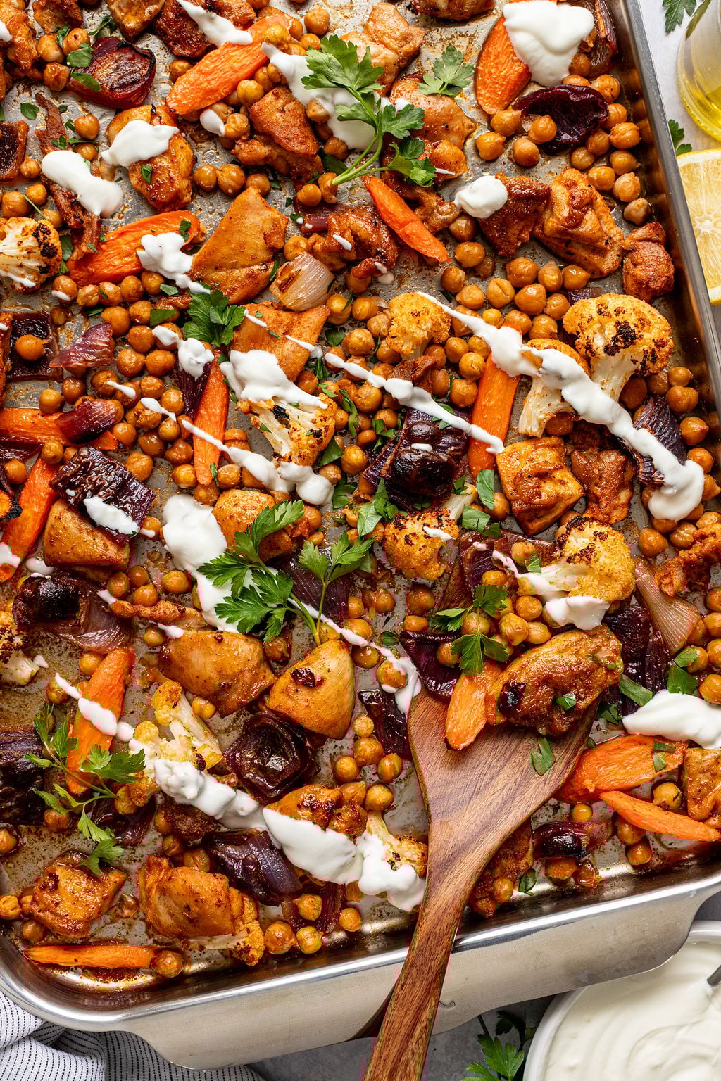 Roasted chickpeas, chicken, and vegetables drizzled with yogurt sauce on a sheet pan.