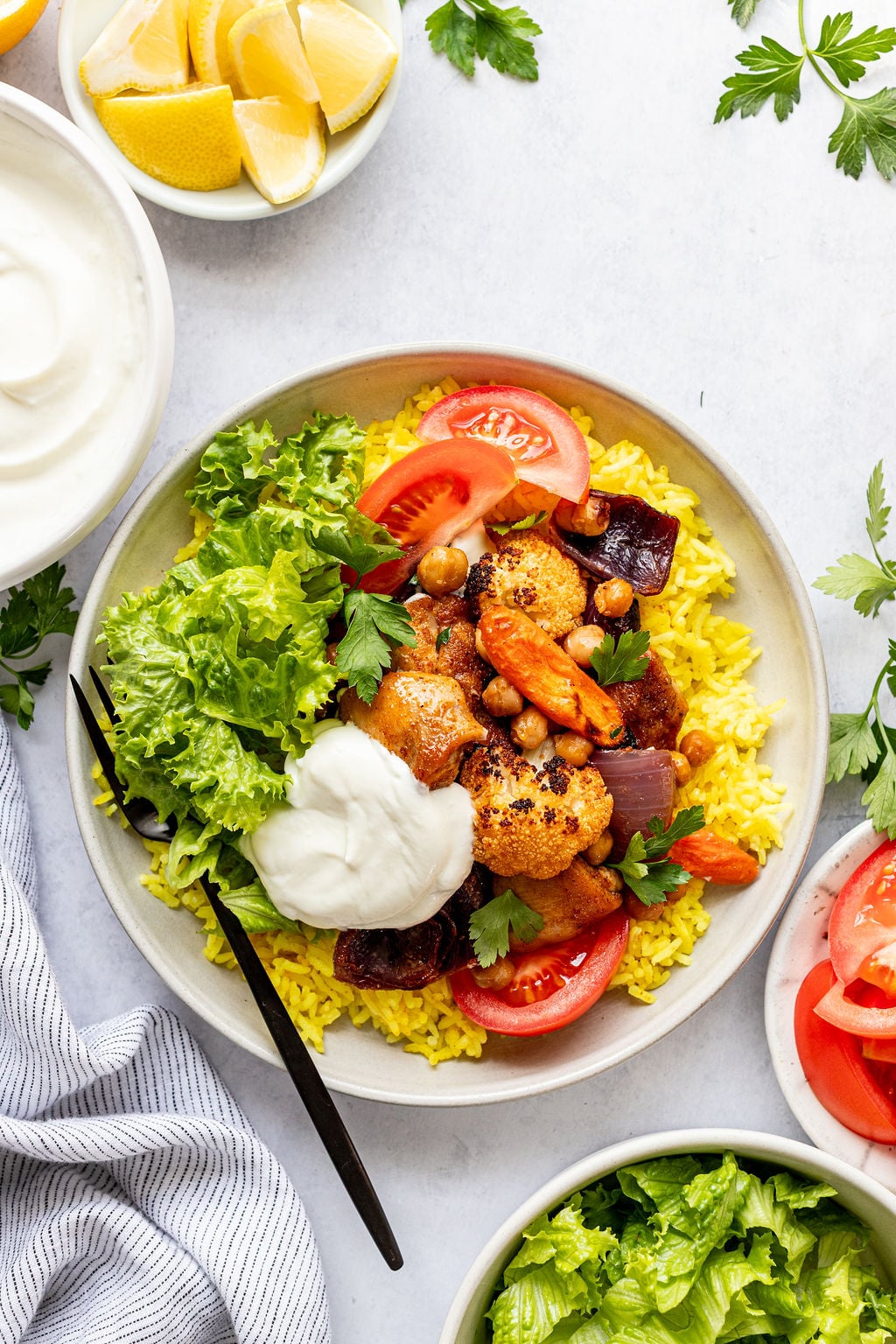Lettuce, tomatoes, yellow rice, yogurt sauce, and roasted chickpeas, chicken, carrots, cauliflower, and red onion on a plate.