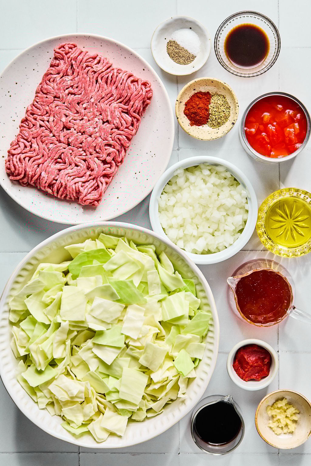 Assorted ingredients for a cabbage and ground beef dish, including raw ground beef, chopped cabbage, diced onions, tomato products, spices, garlic, and liquids, arranged in bowls.