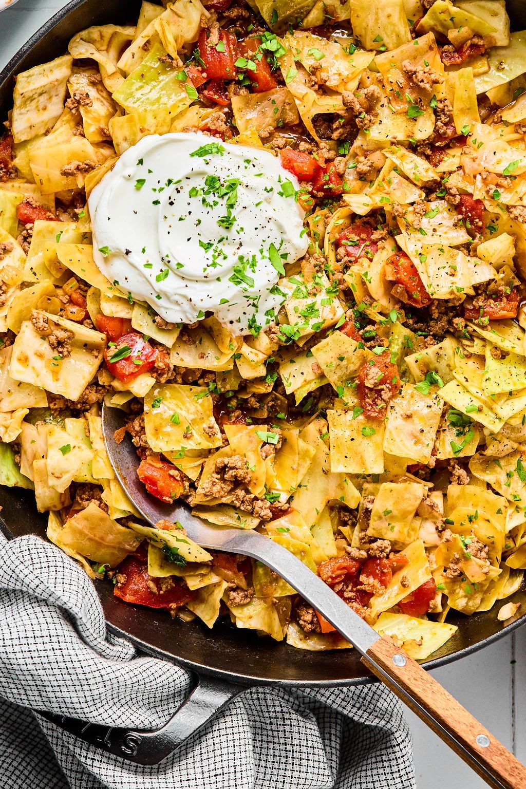 A skillet filled with cooked cabbage, ground meat, and diced tomatoes, topped with a dollop of sour cream and chopped herbs, with a serving spoon.