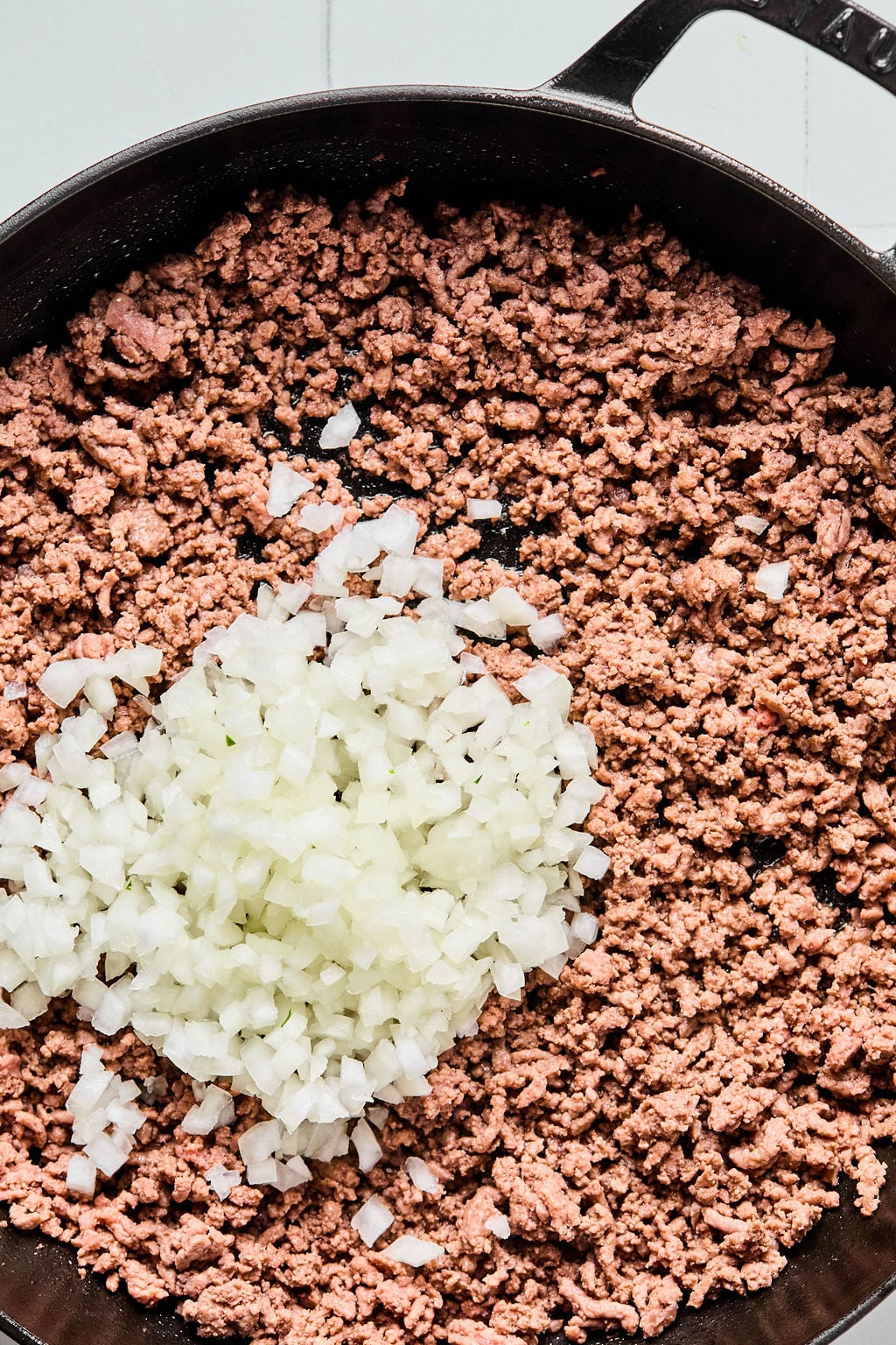 Ground beef and chopped onions cooking in a black skillet on a stovetop.