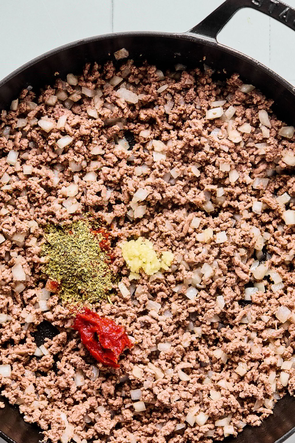 Ground beef and diced onions cooking in a cast iron pan with added tomato paste, minced garlic, and dried herbs.