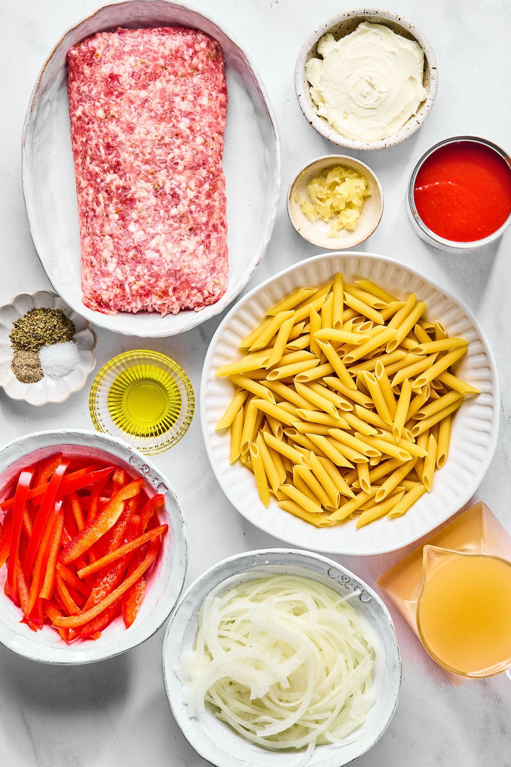A top-down view of penne pasta, ground meat, tomato sauce, sliced red peppers, sliced onions, minced garlic, spices, oil, and broth in separate bowls on a white surface.