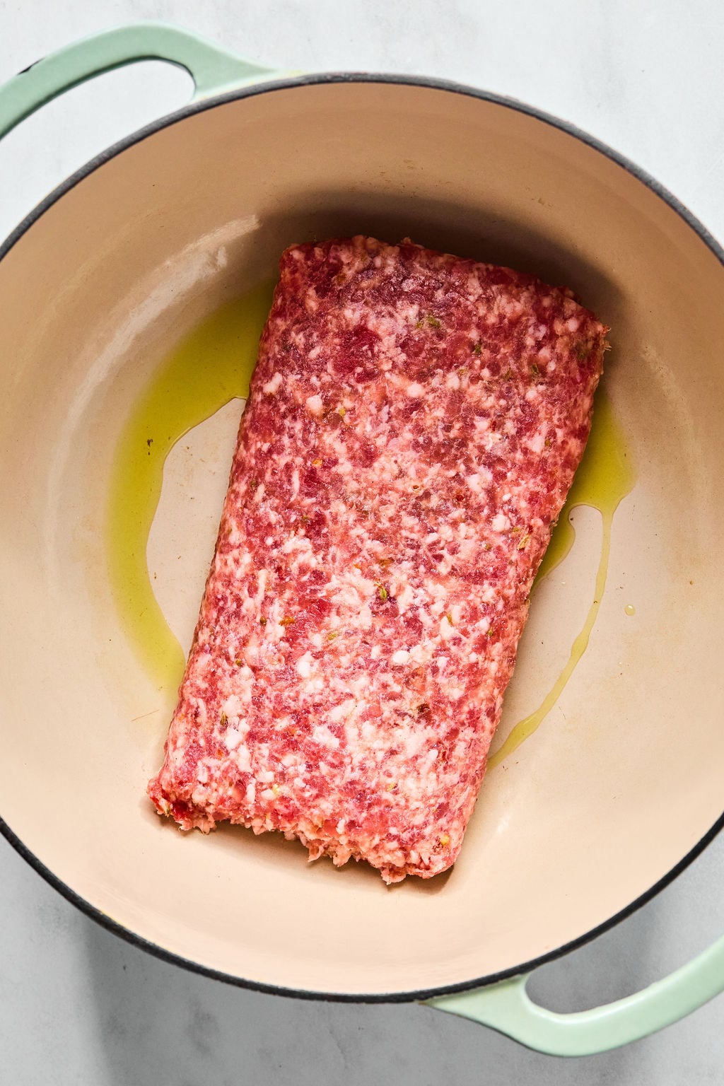 Raw ground meat loaf placed in a large beige pot with a thin layer of olive oil on the bottom, ready for cooking.