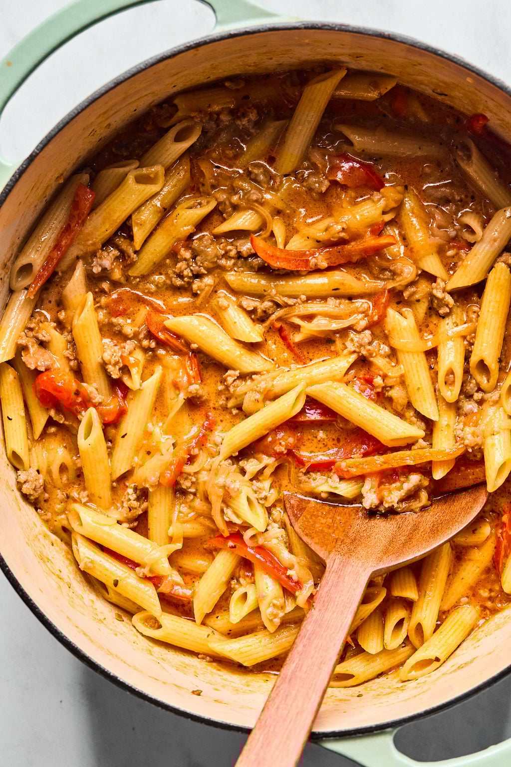 A pot filled with cooked penne pasta, ground meat, sliced tomatoes, and creamy sauce, with a wooden spoon resting inside.