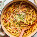 A pot of cooked penne pasta with a tomato-based sauce, ground meat, herbs, and grated cheese, with a wooden spoon resting inside.