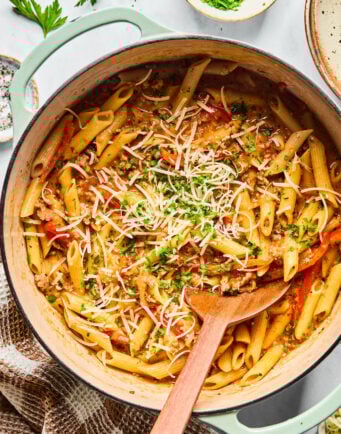 A pot of cooked penne pasta with a tomato-based sauce, ground meat, herbs, and grated cheese, with a wooden spoon resting inside.