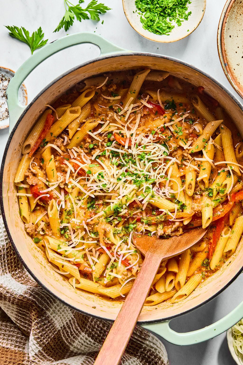 A pot of cooked penne pasta with a tomato-based sauce, ground meat, herbs, and grated cheese, with a wooden spoon resting inside.