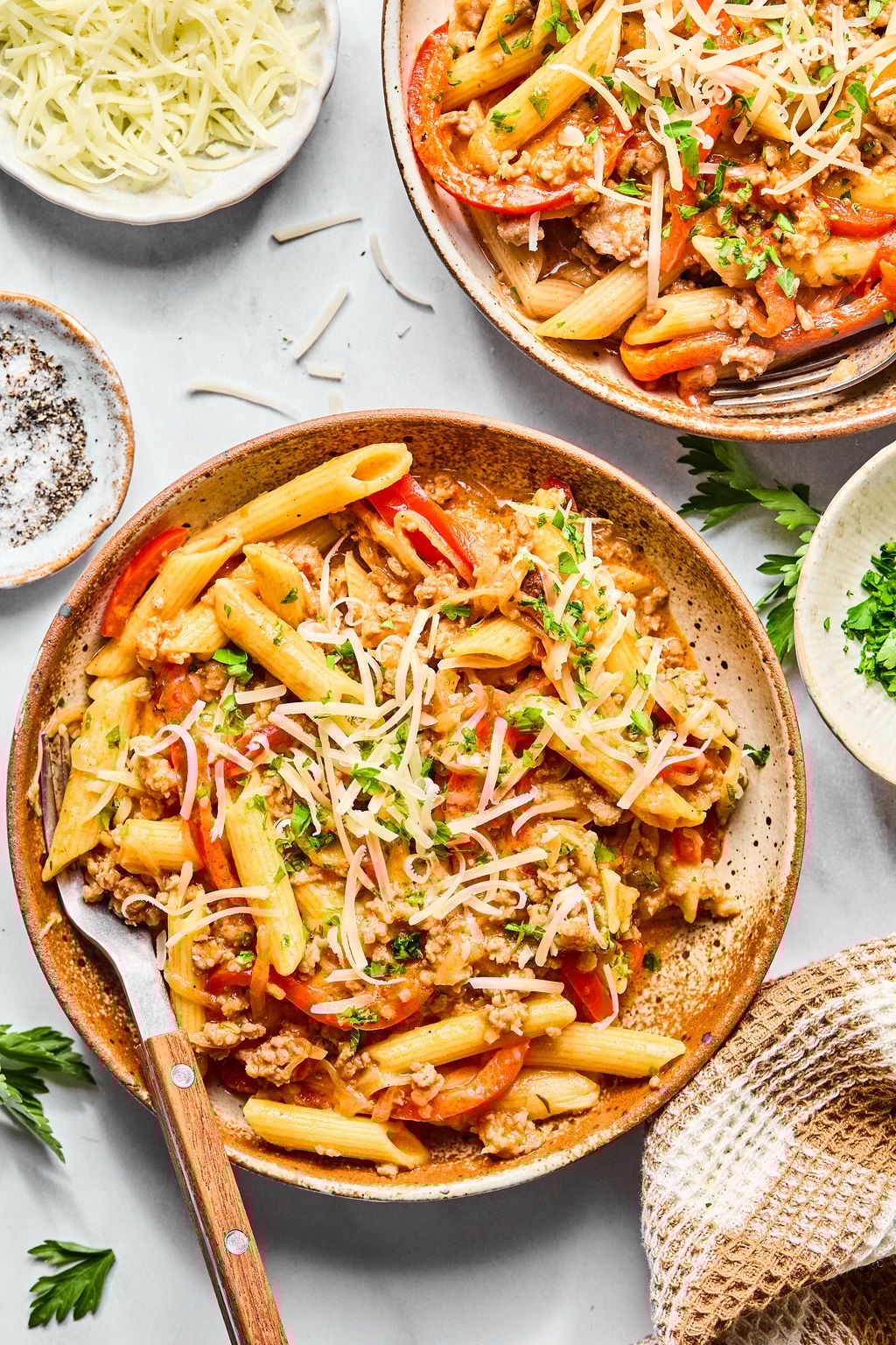 A bowl of penne pasta with ground meat, bell peppers, and shredded cheese, garnished with chopped parsley.