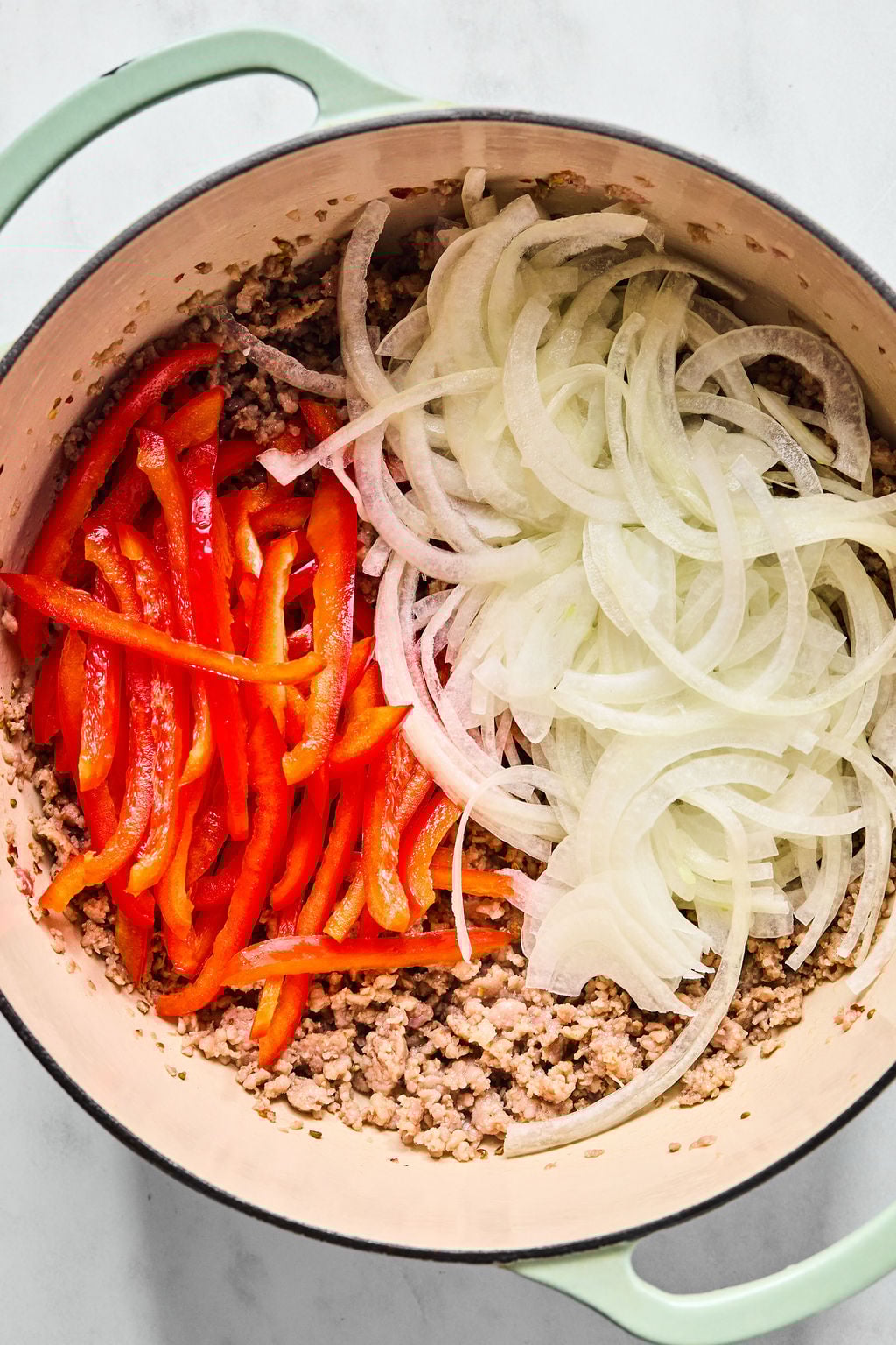 Sliced red bell peppers and onions on top of cooked ground meat in a large pot.