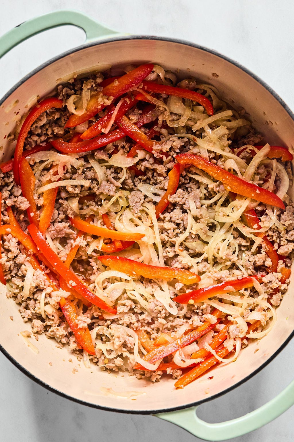 A large pot containing cooked ground meat, sliced onions, and red bell peppers on a light surface.