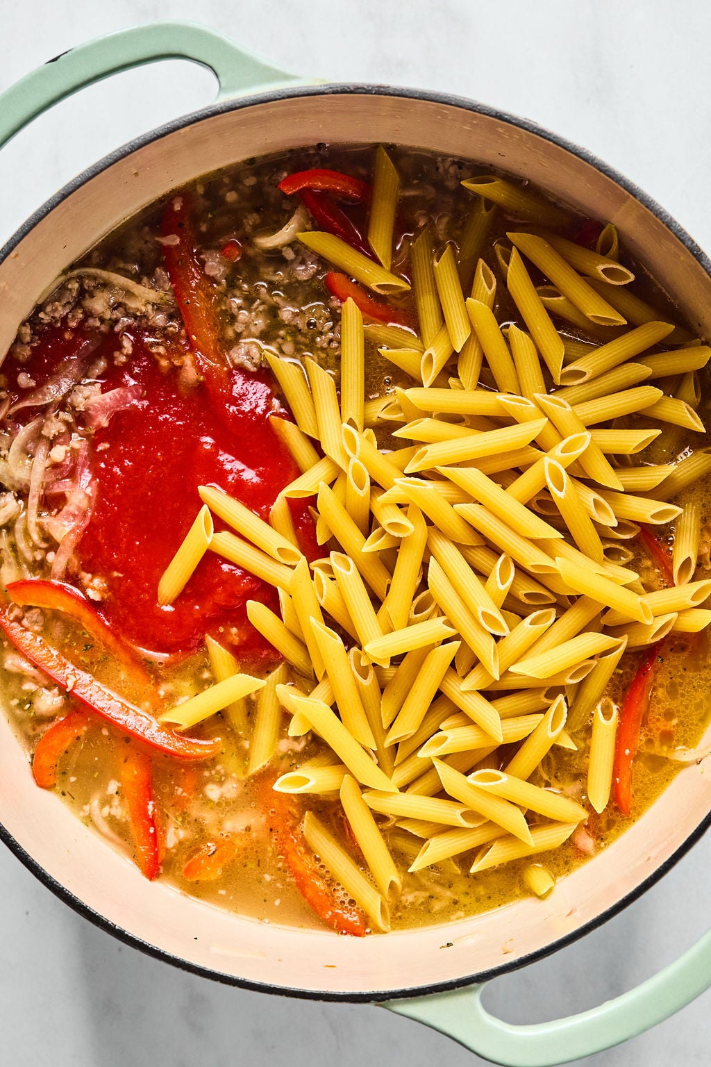 Large pot containing penne pasta, red bell peppers, ground meat, tomato sauce, and broth.