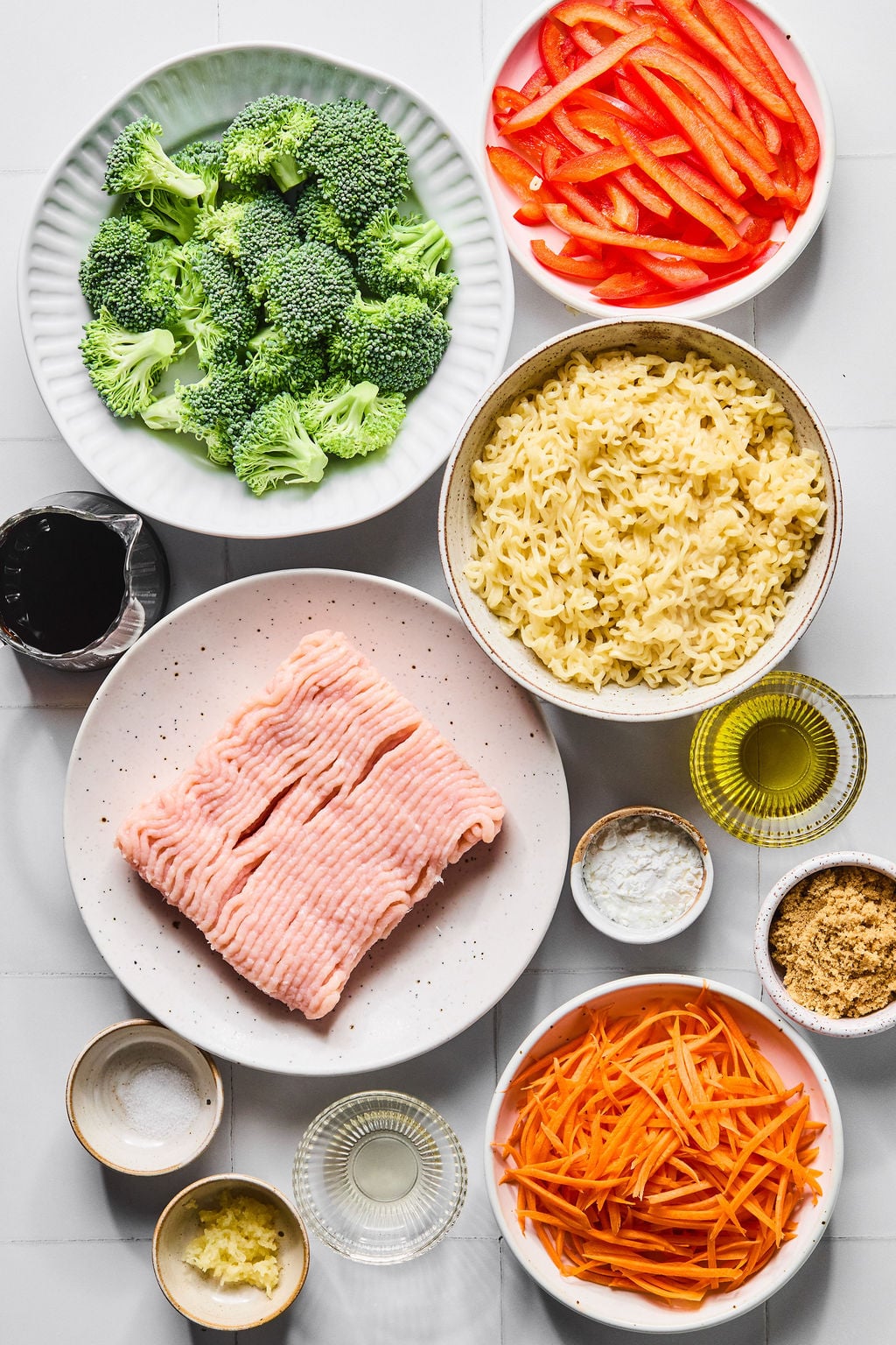 A variety of ingredients in bowls, including broccoli, red bell pepper, cooked noodles, ground turkey, carrots, garlic, sauces, oil, and seasonings, arranged on a white surface.
