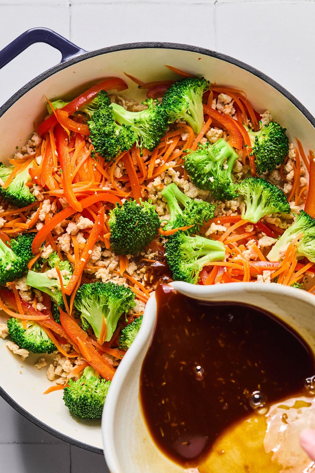 A pot with saut&eacute;ed broccoli, shredded carrots, and ground meat, with a bowl of dark sauce being poured in.