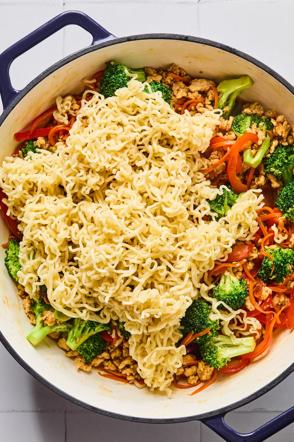 A pot filled with cooked noodles, broccoli, red bell peppers, carrots, and ground chicken, ready to be mixed together.