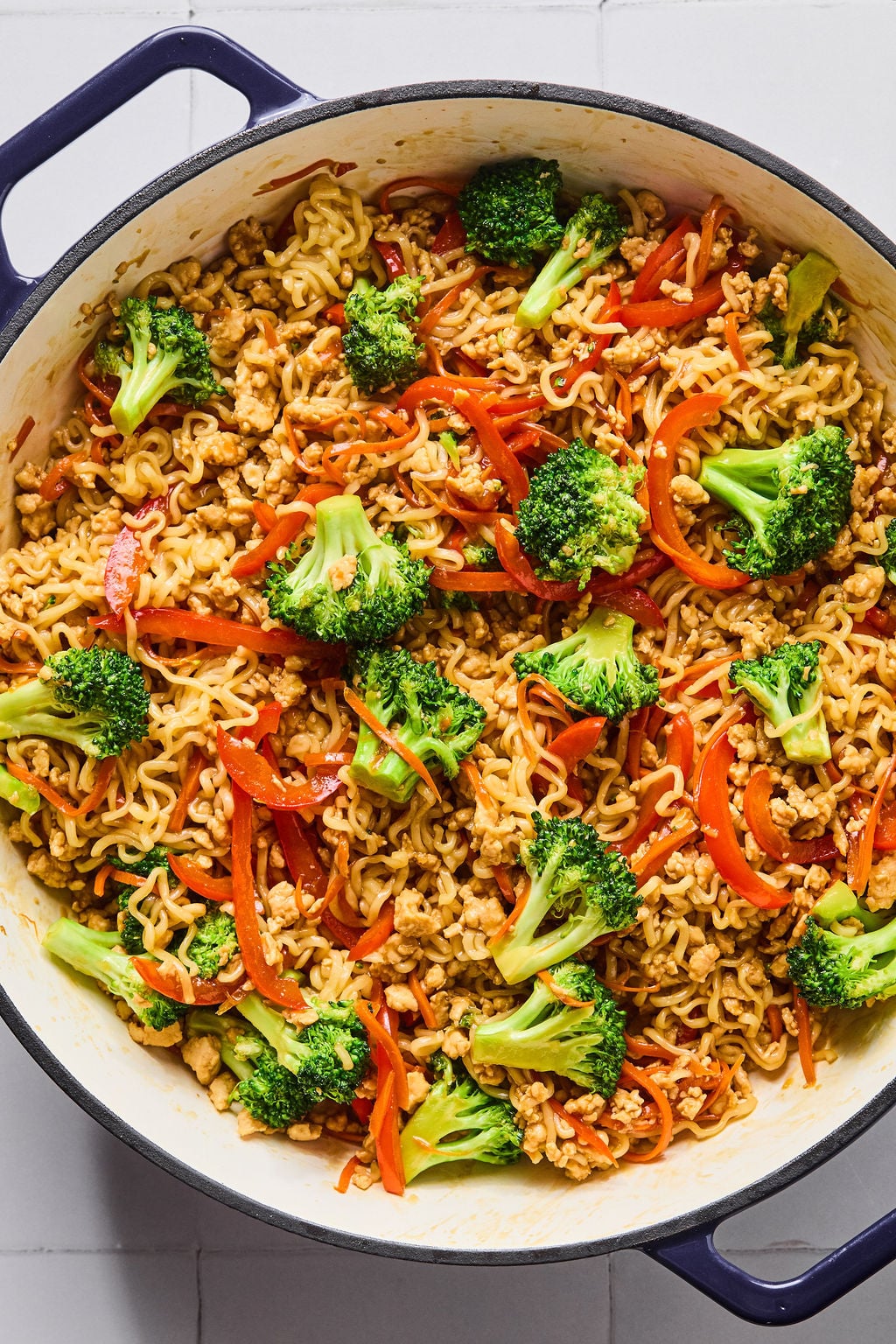 A large pot filled with cooked ramen noodles, broccoli florets, red bell pepper strips, and ground chicken mixed together.