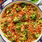 A large pot filled with cooked ramen noodles, broccoli, red bell peppers, and green onions, garnished with sesame seeds, sits on a white countertop with bowls of toppings nearby.