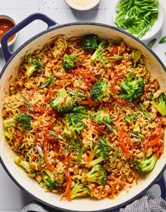A large pot filled with cooked ramen noodles, broccoli, red bell peppers, and green onions, garnished with sesame seeds, sits on a white countertop with bowls of toppings nearby.