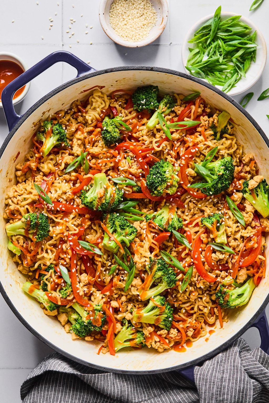 A large pot filled with cooked ramen noodles, broccoli, red bell peppers, and green onions, garnished with sesame seeds, sits on a white countertop with bowls of toppings nearby.