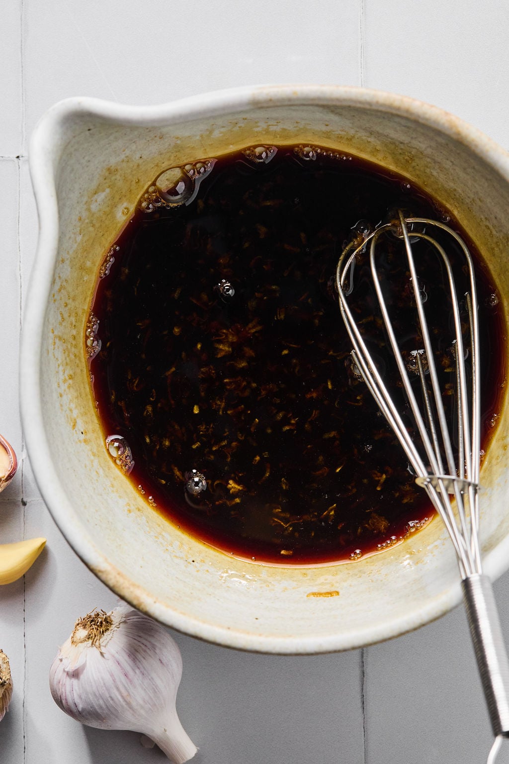 A ceramic bowl filled with dark marinade and minced garlic, with a metal whisk resting inside.