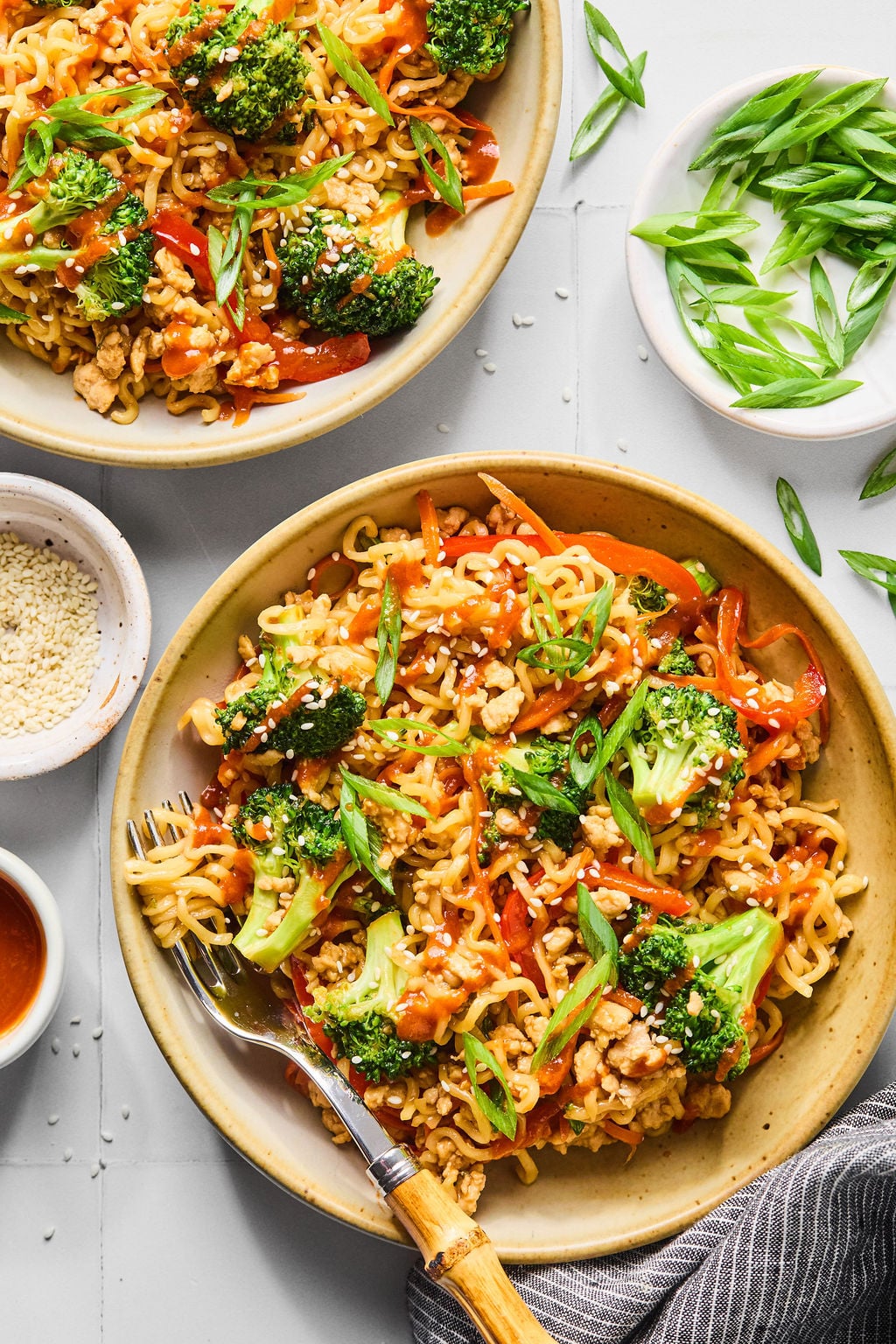 Two plates of stir-fried noodles with broccoli, bell peppers, carrots, chicken, and green onions, garnished with sesame seeds.