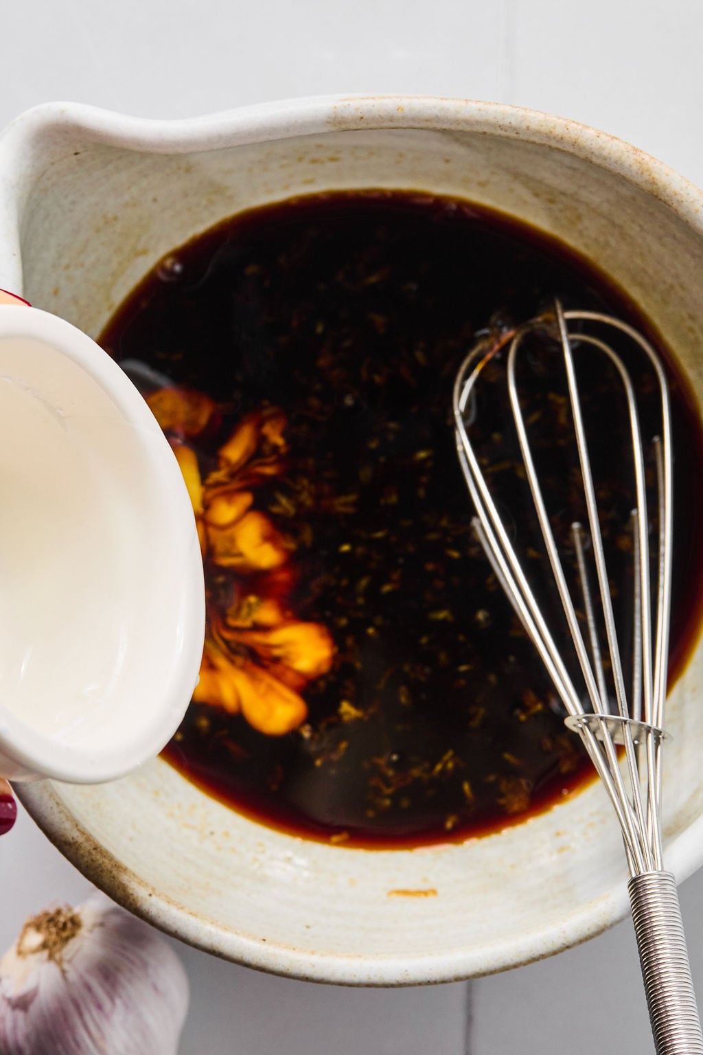 A person pours a white liquid from a small white pitcher into a bowl of dark sauce.