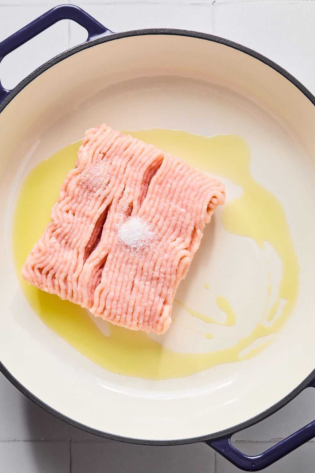 Raw ground turkey with salt is placed in a white pan with a layer of olive oil, ready for cooking.