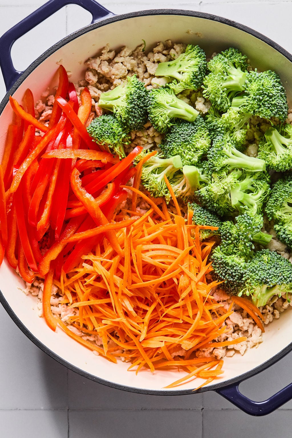 A pot containing raw sliced red bell peppers, shredded carrots, broccoli florets, and cooked ground meat on a white tile surface.