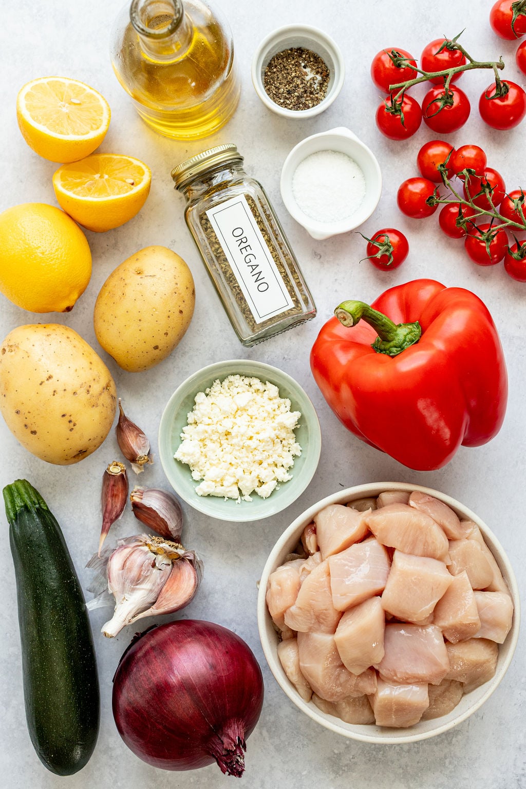 Top-down view of raw ingredients including diced chicken, red onion, zucchini, garlic, potatoes, lemon, cherry tomatoes, red bell pepper, feta cheese, oregano, olive oil, salt, and pepper.