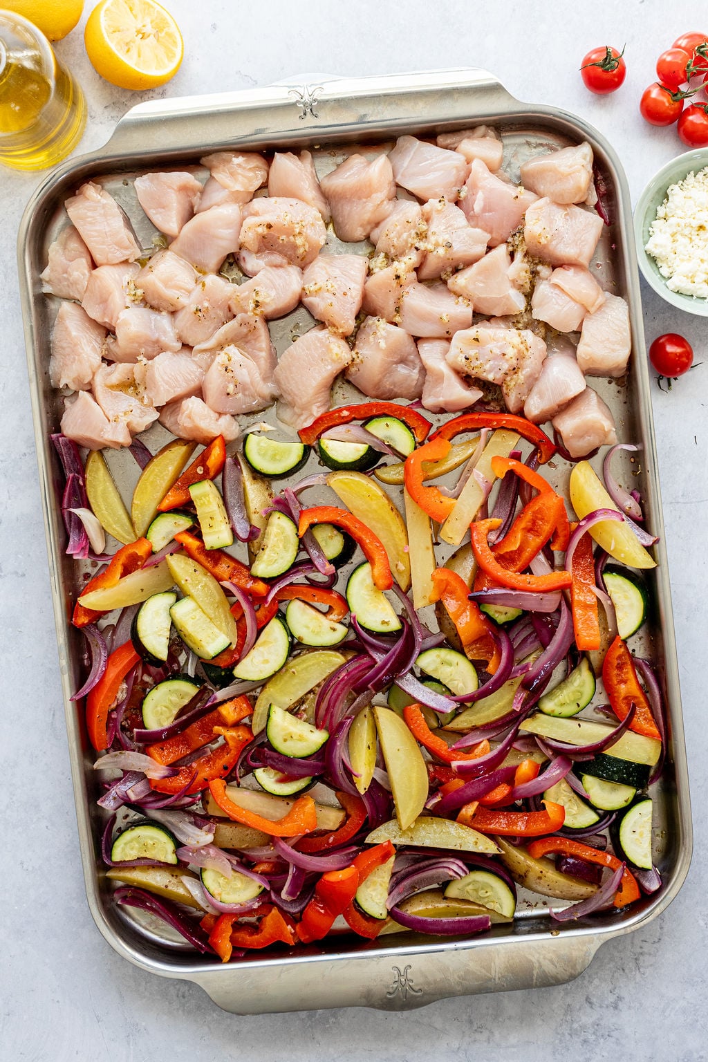Sheet pan with raw diced chicken on one side and sliced zucchini, bell peppers, potatoes, and red onions on the other side.