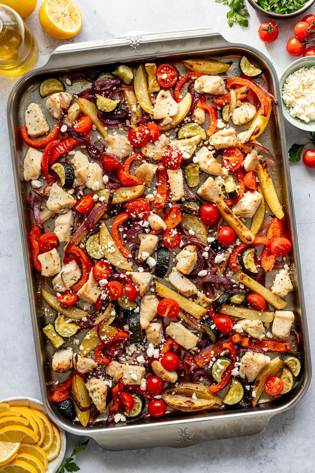 Sheet pan with cooked chicken pieces, sliced bell peppers, zucchini, potatoes, cherry tomatoes, red onion, and crumbled cheese, garnished with lemon and herbs.