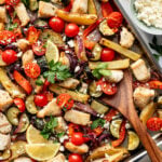 Sheet pan with baked chicken, sliced bell peppers, cherry tomatoes, potatoes, zucchini, red onion, lemon slices, herbs, and crumbled cheese.