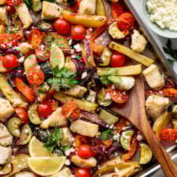 Sheet pan with baked chicken, sliced bell peppers, cherry tomatoes, potatoes, zucchini, red onion, lemon slices, herbs, and crumbled cheese.