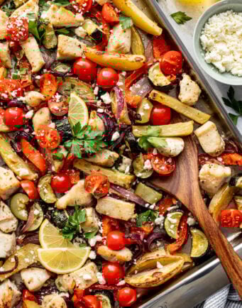 Sheet pan with baked chicken, sliced bell peppers, cherry tomatoes, potatoes, zucchini, red onion, lemon slices, herbs, and crumbled cheese.