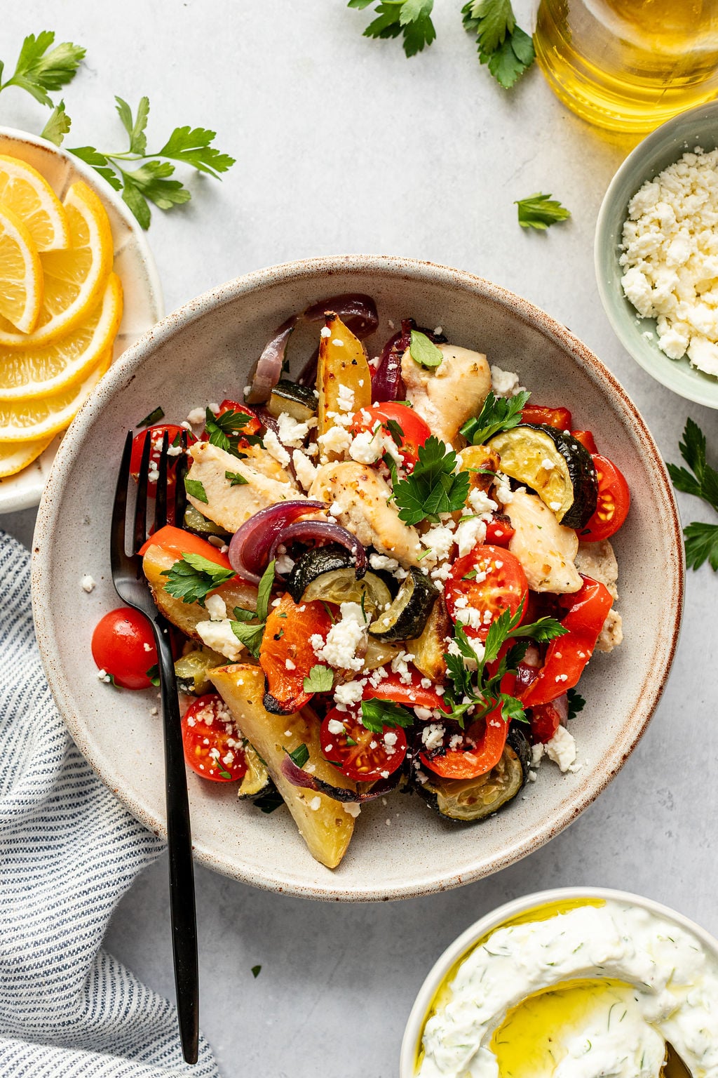 A bowl of roasted chicken and vegetables, topped with crumbled feta and parsley, is served with lemon slices, dip, and olive oil on the side.