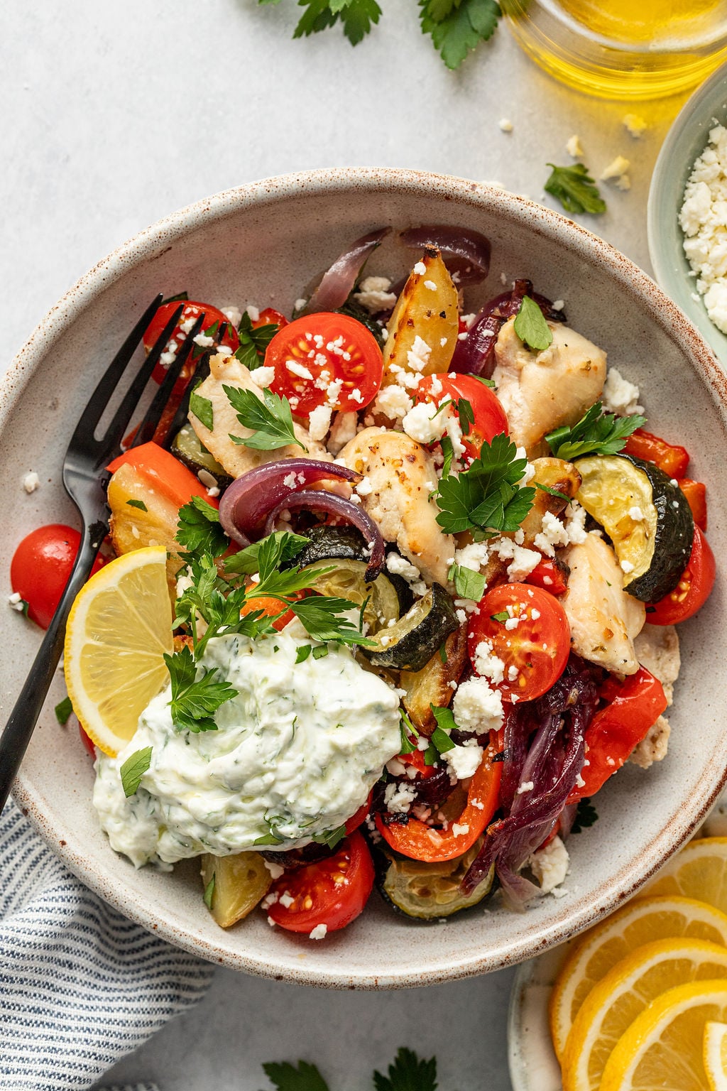 A bowl of Greek chicken with roasted vegetables, cherry tomatoes, tzatziki, feta cheese, fresh parsley, and a lemon wedge, served with a fork.