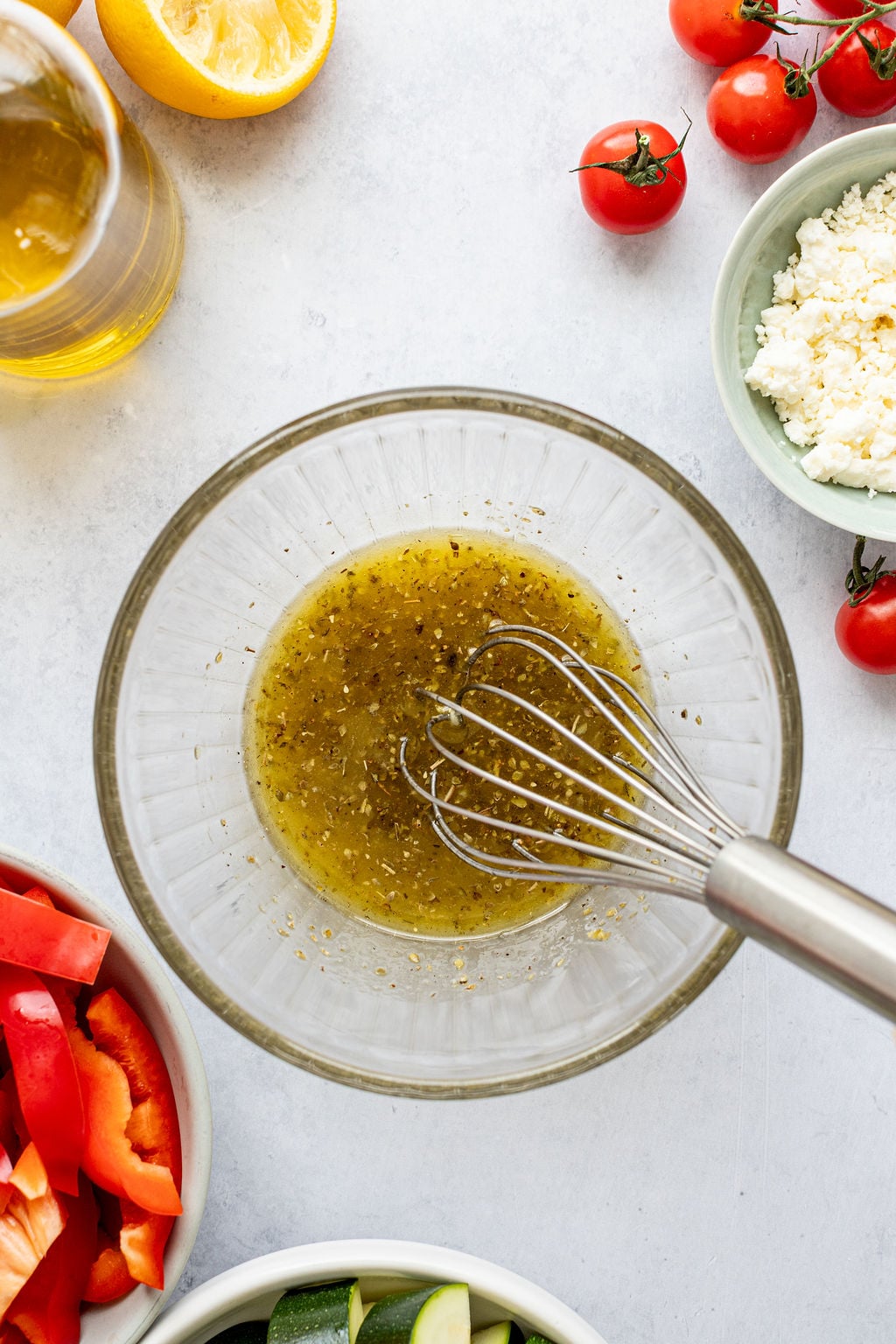 A mixing bowl with marinade and a whisk, surrounded by chopped vegetables, feta cheese, tomatoes, a lemon half, and a glass of olive oil on a white surface.