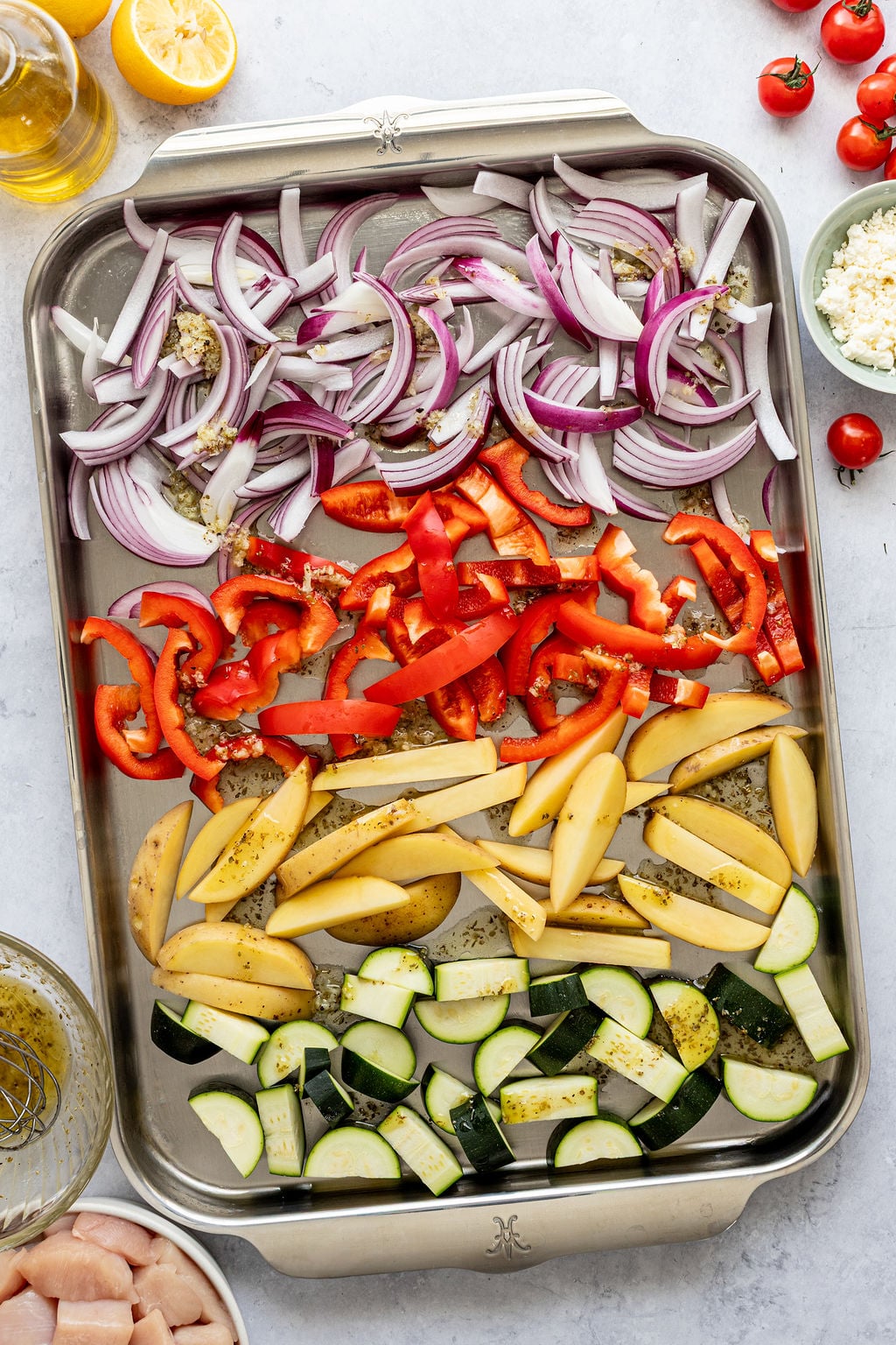 A baking tray with rows of sliced red onion, red bell pepper, yellow potato, and zucchini, seasoned with herbs, ready to be roasted.