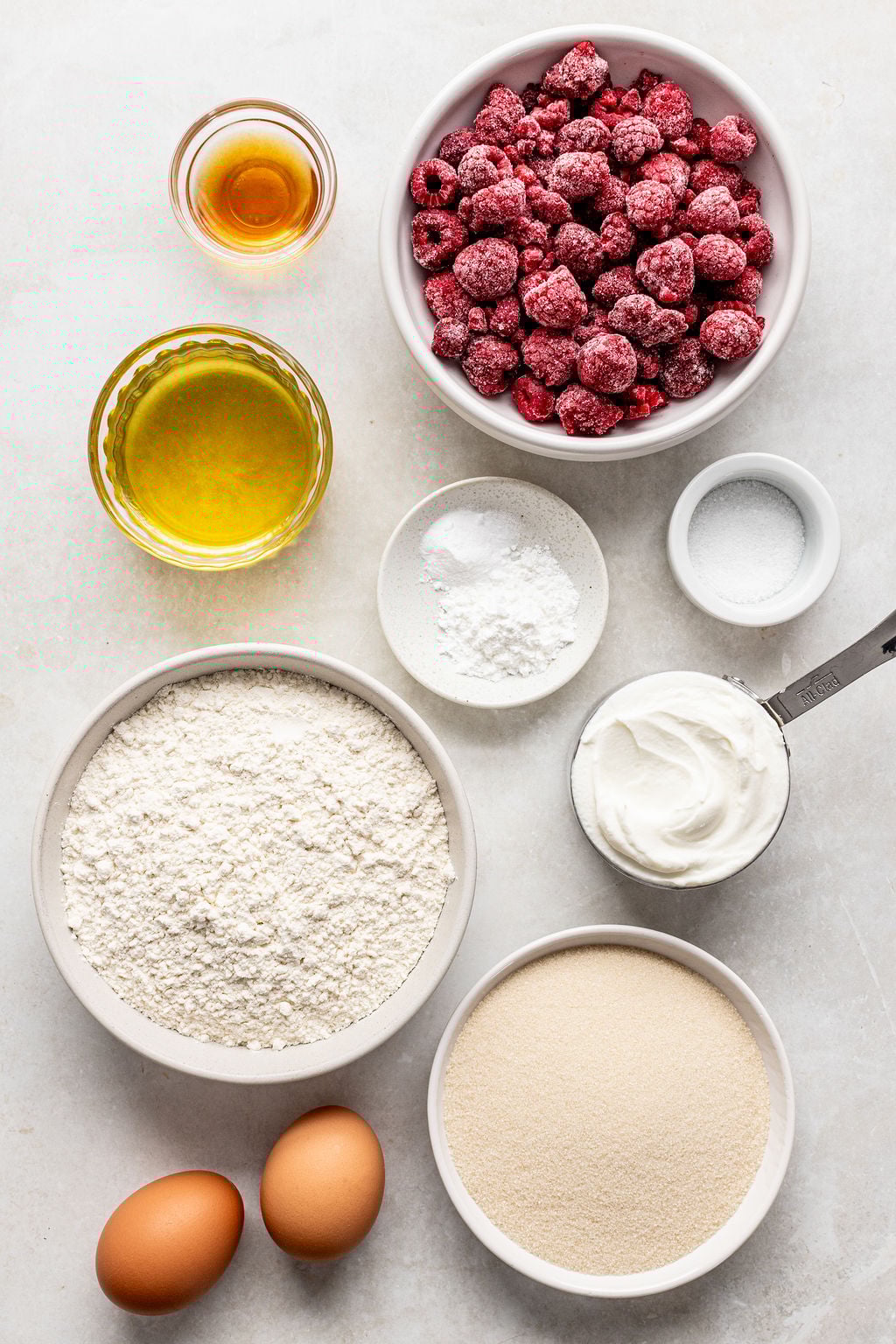 A flat lay of baking ingredients including frozen raspberries, flour, sugar, eggs, oil, vanilla extract, yogurt, baking powder, and salt, arranged in bowls on a white surface.
