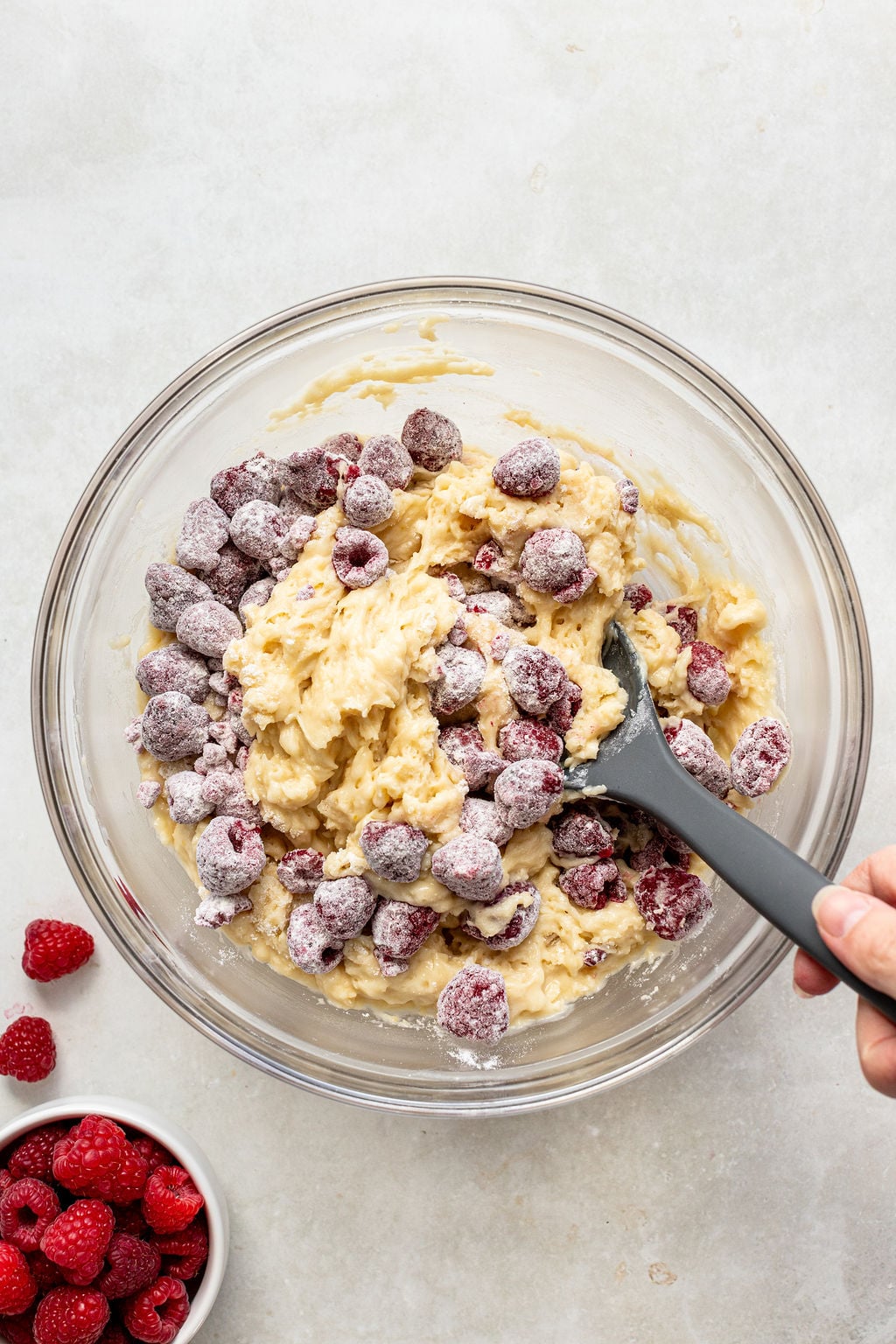 A hand stirs muffin batter with raspberries in a glass bowl using a black spatula; a small bowl of fresh raspberries sits nearby.