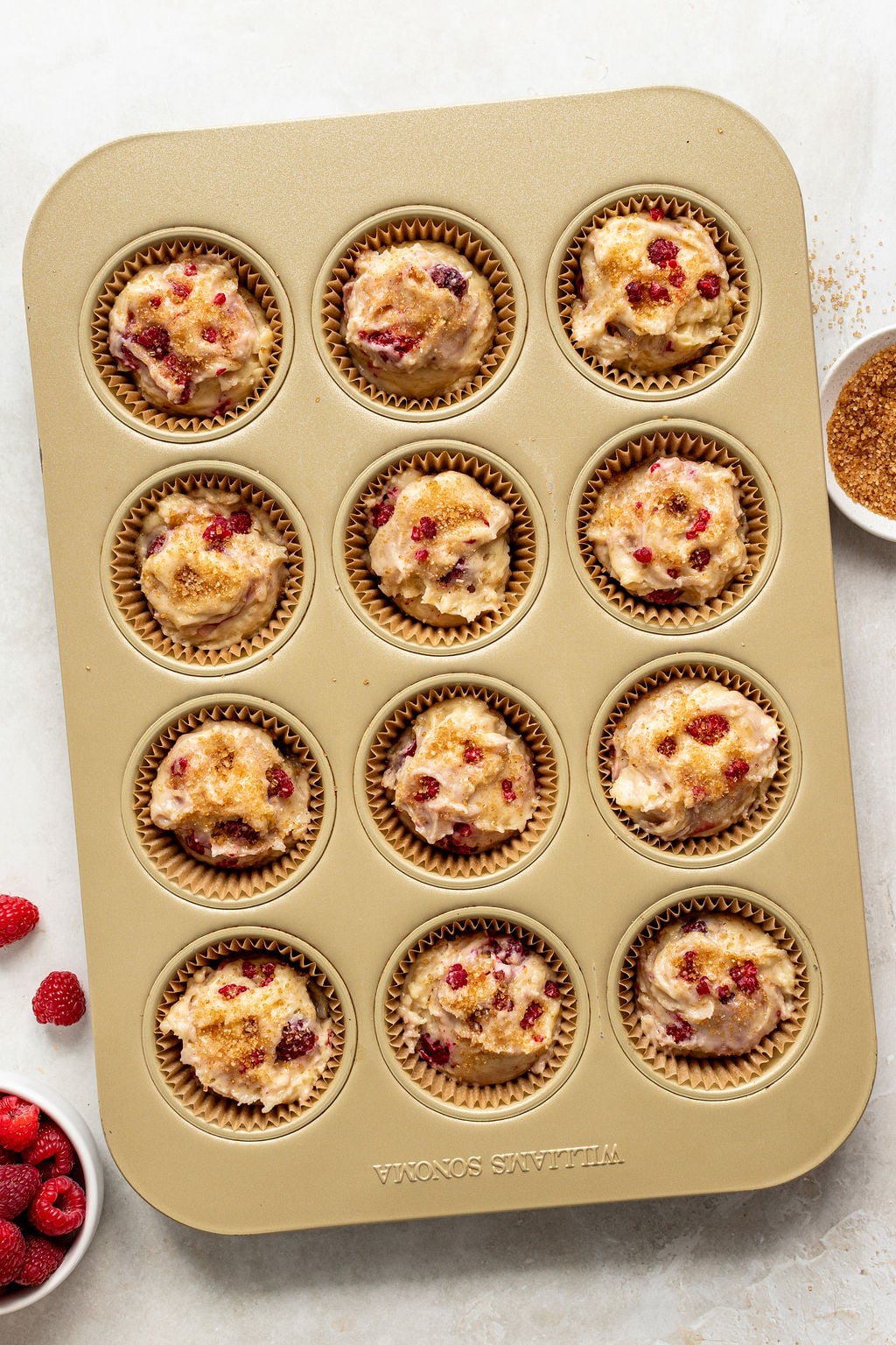 A muffin tin filled with unbaked raspberry muffin batter in paper liners, with raspberries and brown sugar in small bowls nearby.