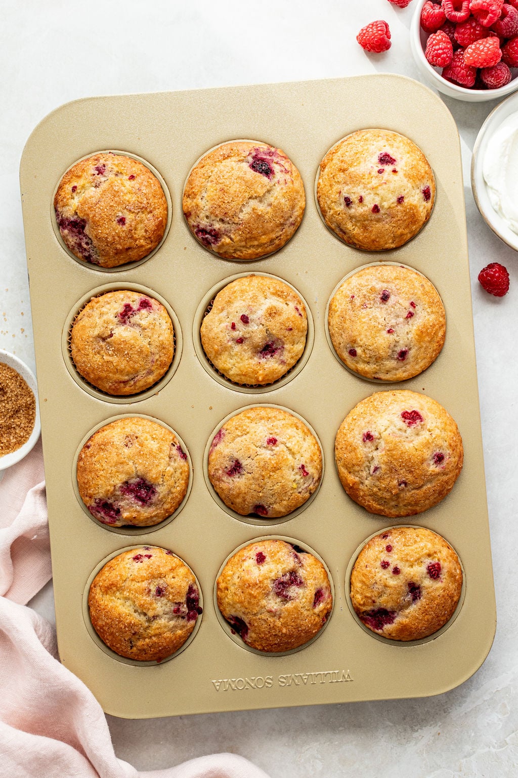 A muffin tin filled with twelve golden brown raspberry muffins, with fresh raspberries and a bowl of cream nearby.