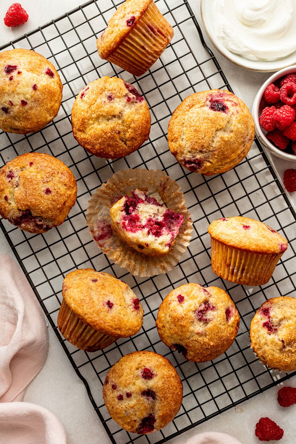 A cooling rack holds several raspberry muffins, one cut open to show the inside. Bowls of raspberries and whipped topping are nearby on a white surface.