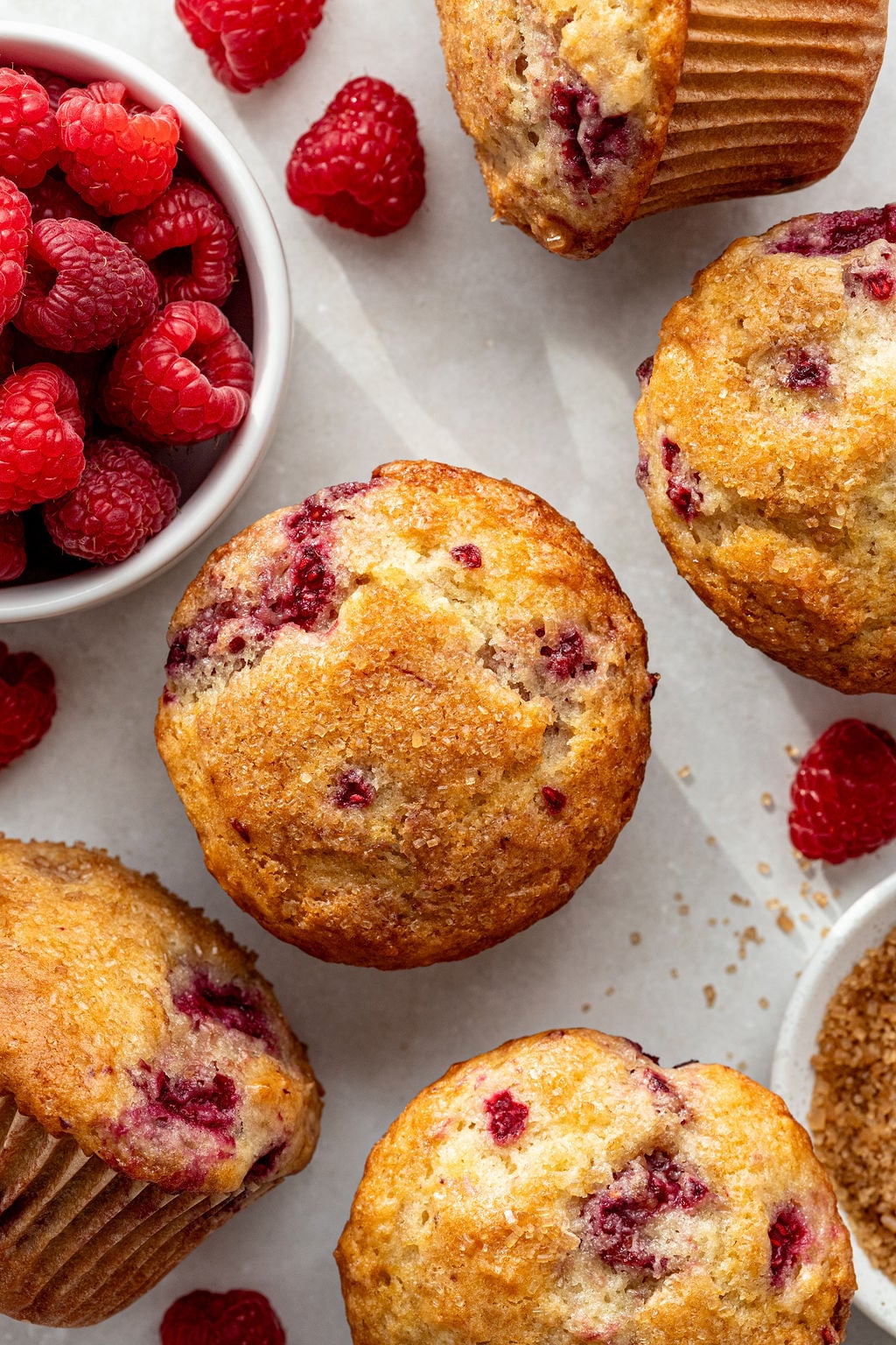Five raspberry muffins are arranged on a light surface next to a small bowl of fresh raspberries and some scattered brown sugar.