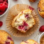 A partially eaten raspberry muffin sits on an open wrapper, surrounded by whole muffins and fresh raspberries on a light surface.